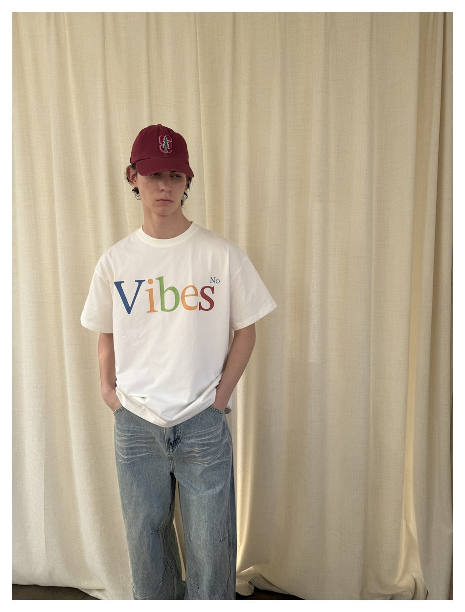 Model wearing a white T-shirt with multicolored “Vibes No” lettering, blue jeans, and a burgundy cap.