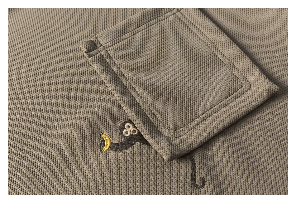 Close-up of a taupe textured garment with a stitched patch pocket and small embroidered motifs.