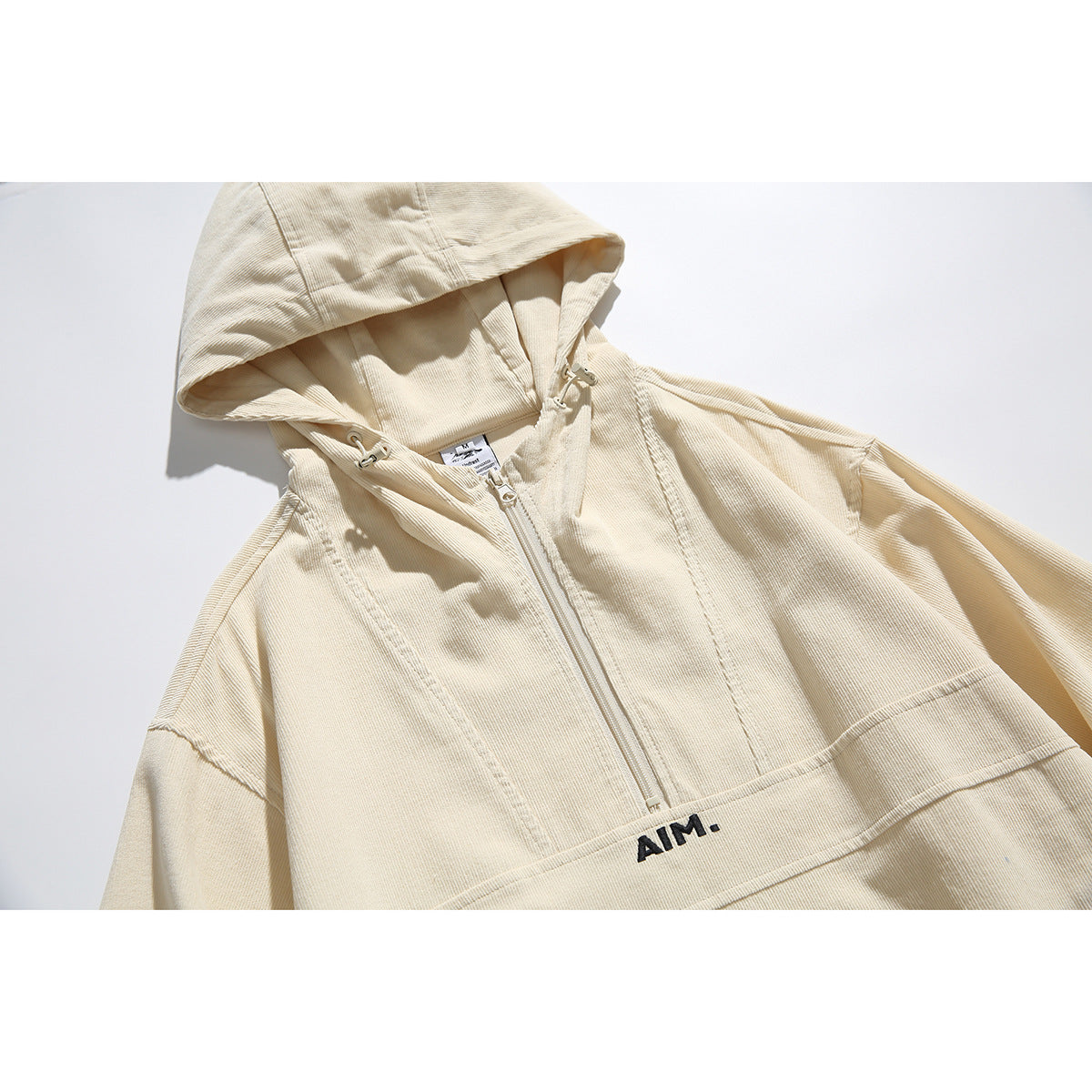 Close-up of a cream hooded pullover jacket showing the hood, front zipper, seams, and small black chest logo.