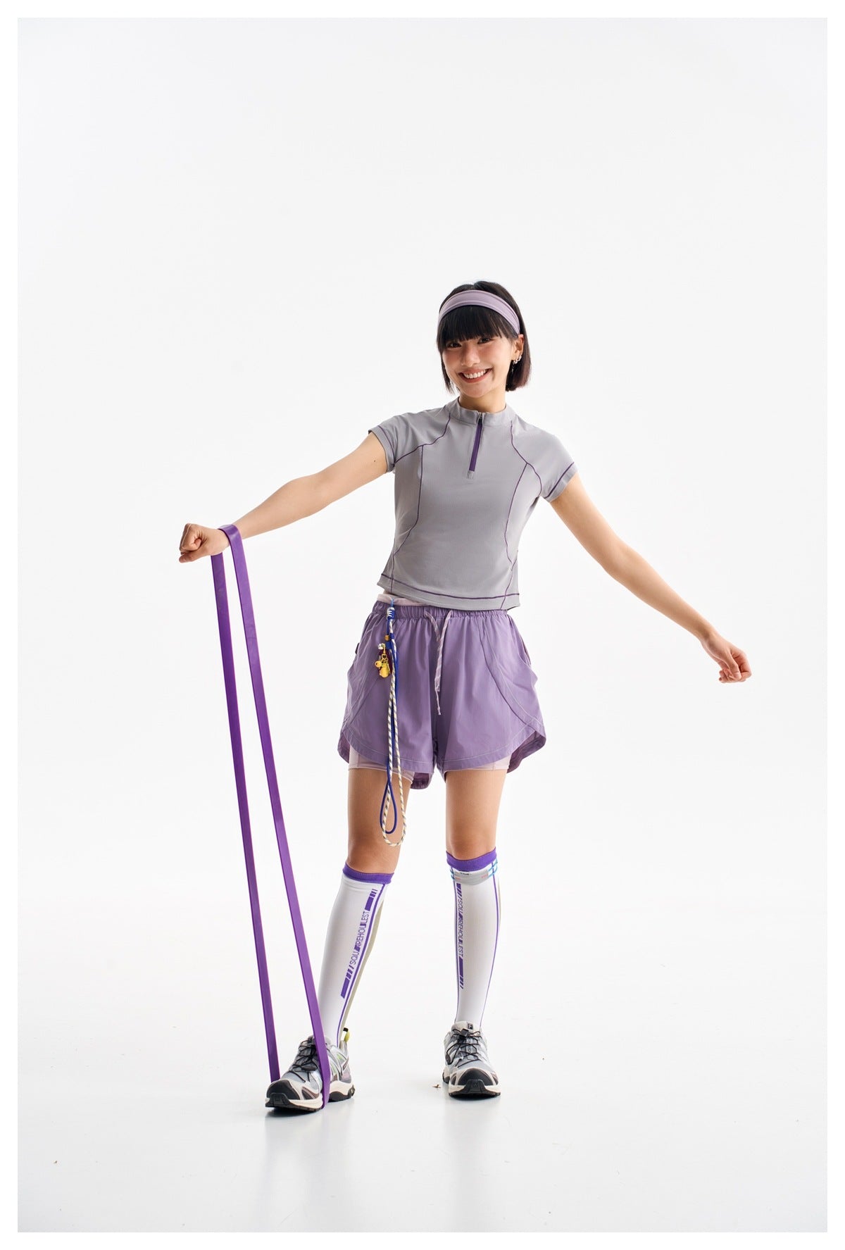 Model in a gray-lilac quarter-zip top and purple shorts, holding a long purple resistance band.