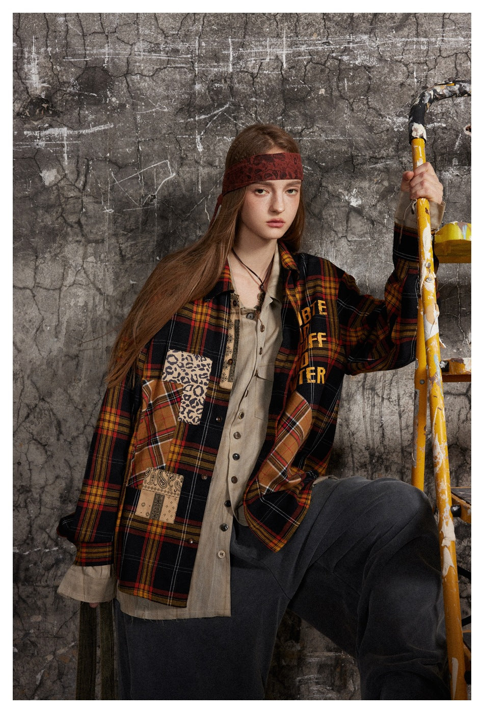 Front view of a model in an oversized plaid patchwork shirt with chest lettering, layered over a beige top.