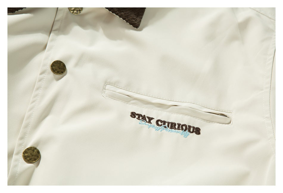 Close-up of an ivory garment with antique-gold buttons, a dark brown collar edge, and “STAY CURIOUS” embroidery.