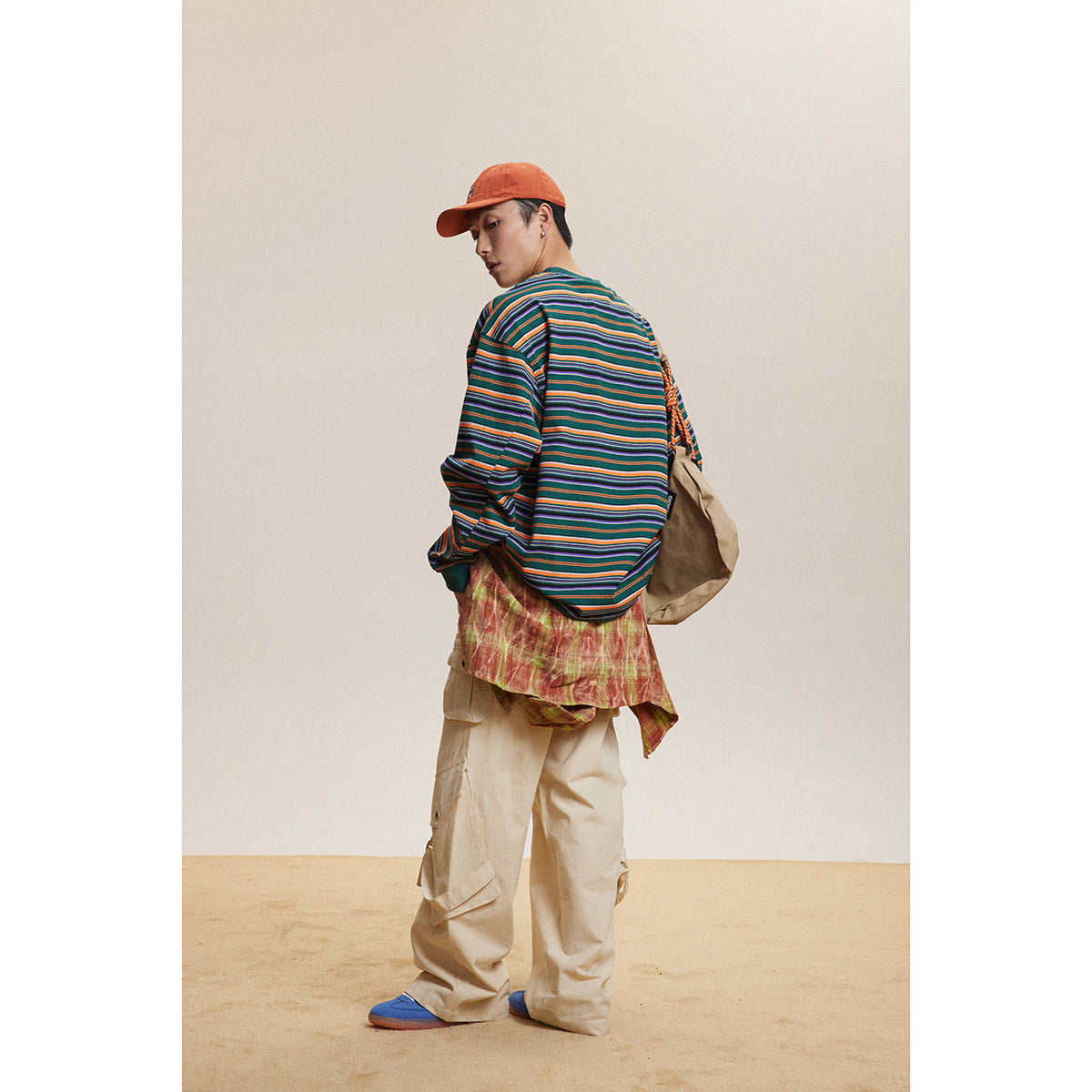 Rear view of a model in a multicolored striped shirt and beige cargo pants, with an orange cap and shoulder bag.