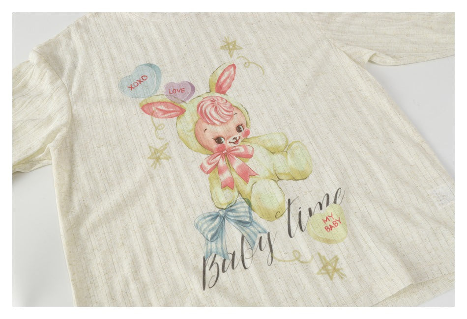 Ivory short-sleeve shirt laid flat with a pastel bunny, pink and blue bows, stars and English lettering.