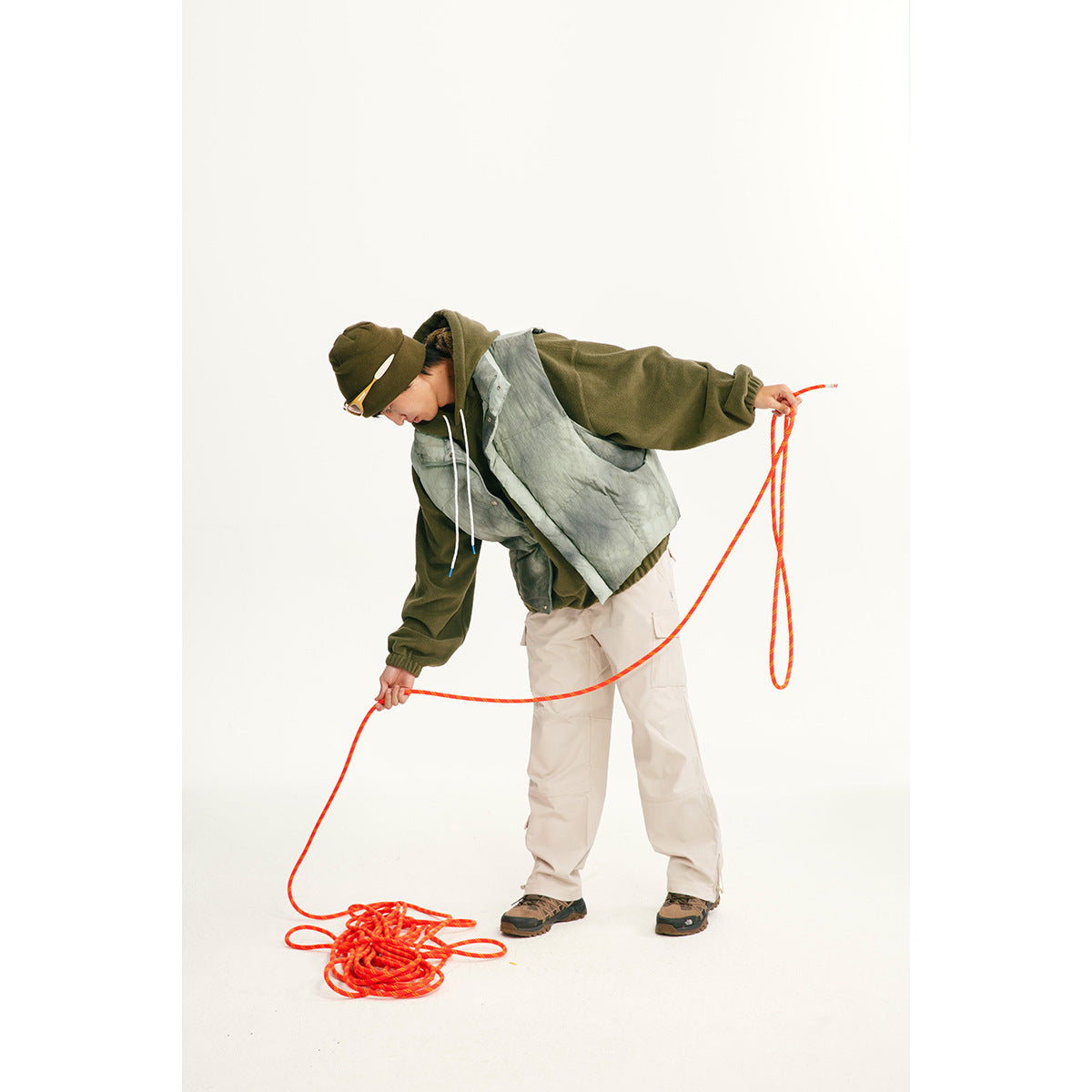 Model wearing a pale gray-green vest over a dark hoodie with light cargo pants, holding a bright orange rope.