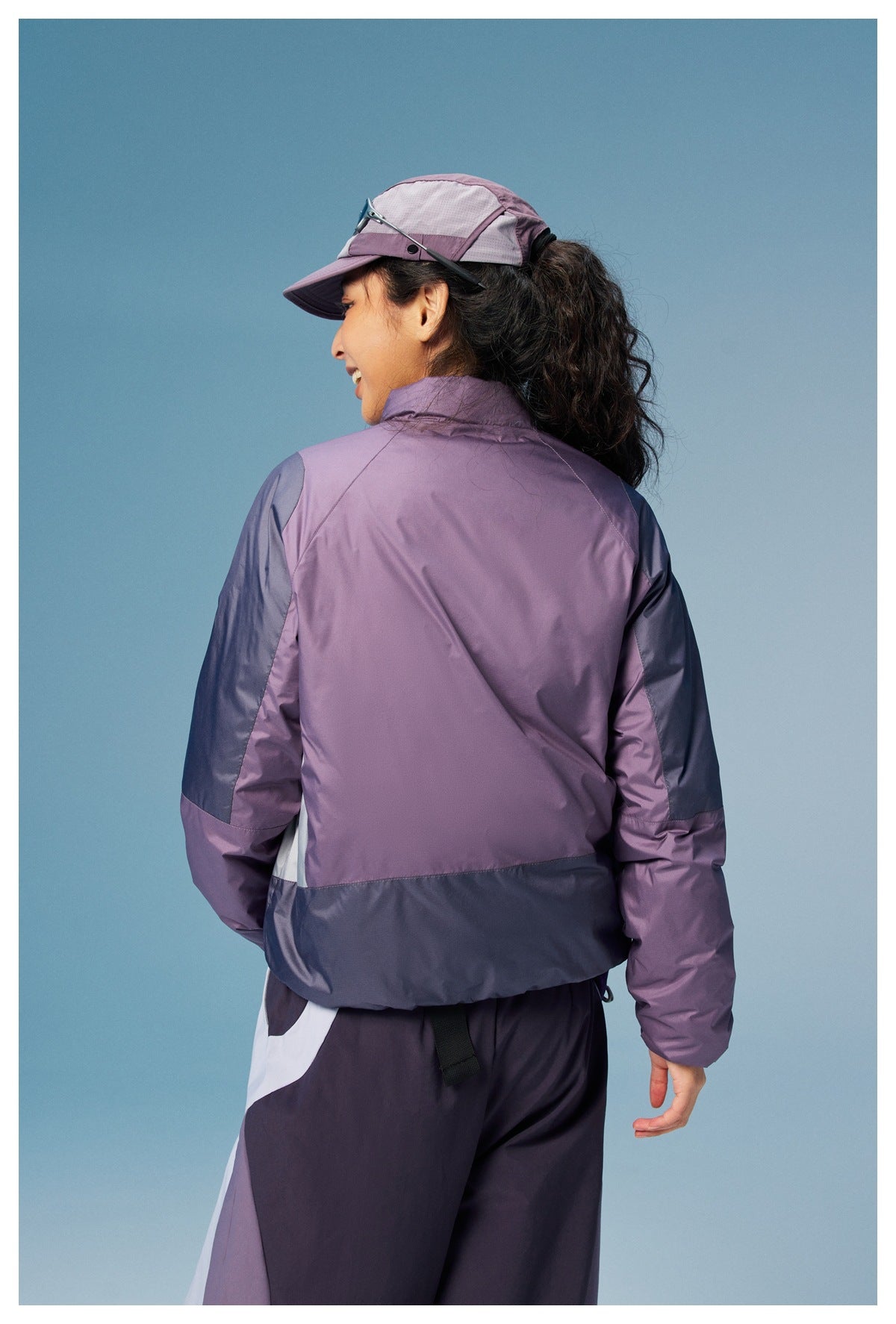 Back view of a model wearing a lavender jacket with charcoal sleeves and hem, paired with color-block pants.