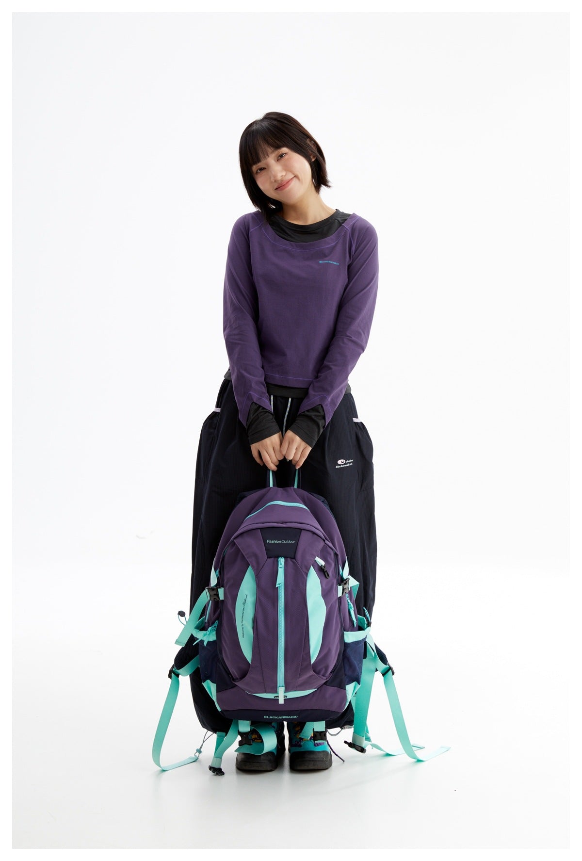 Model wearing a purple long-sleeve top over a black shirt, holding a purple and turquoise hiking backpack.