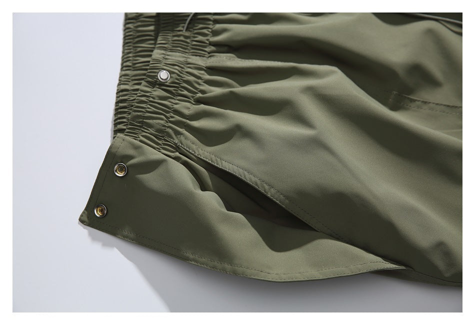 Close-up of olive-green pants showing a gathered elastic edge, stitched fabric and metal snap buttons.