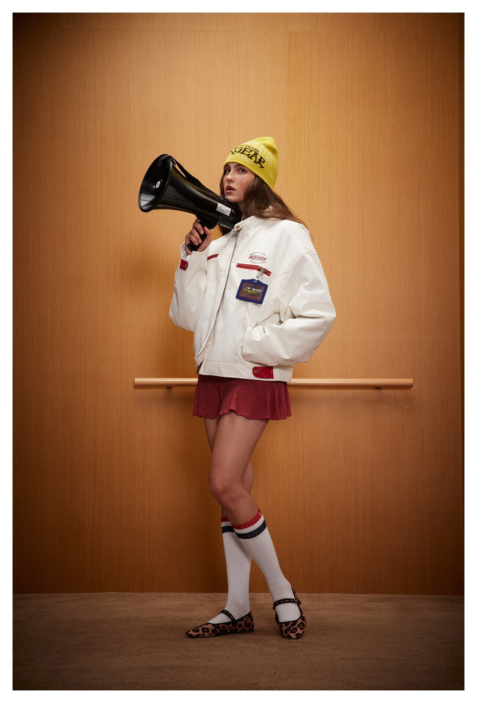 Model wearing a white patch-decorated jacket, red skirt, yellow beanie, and striped socks while holding a megaphone.