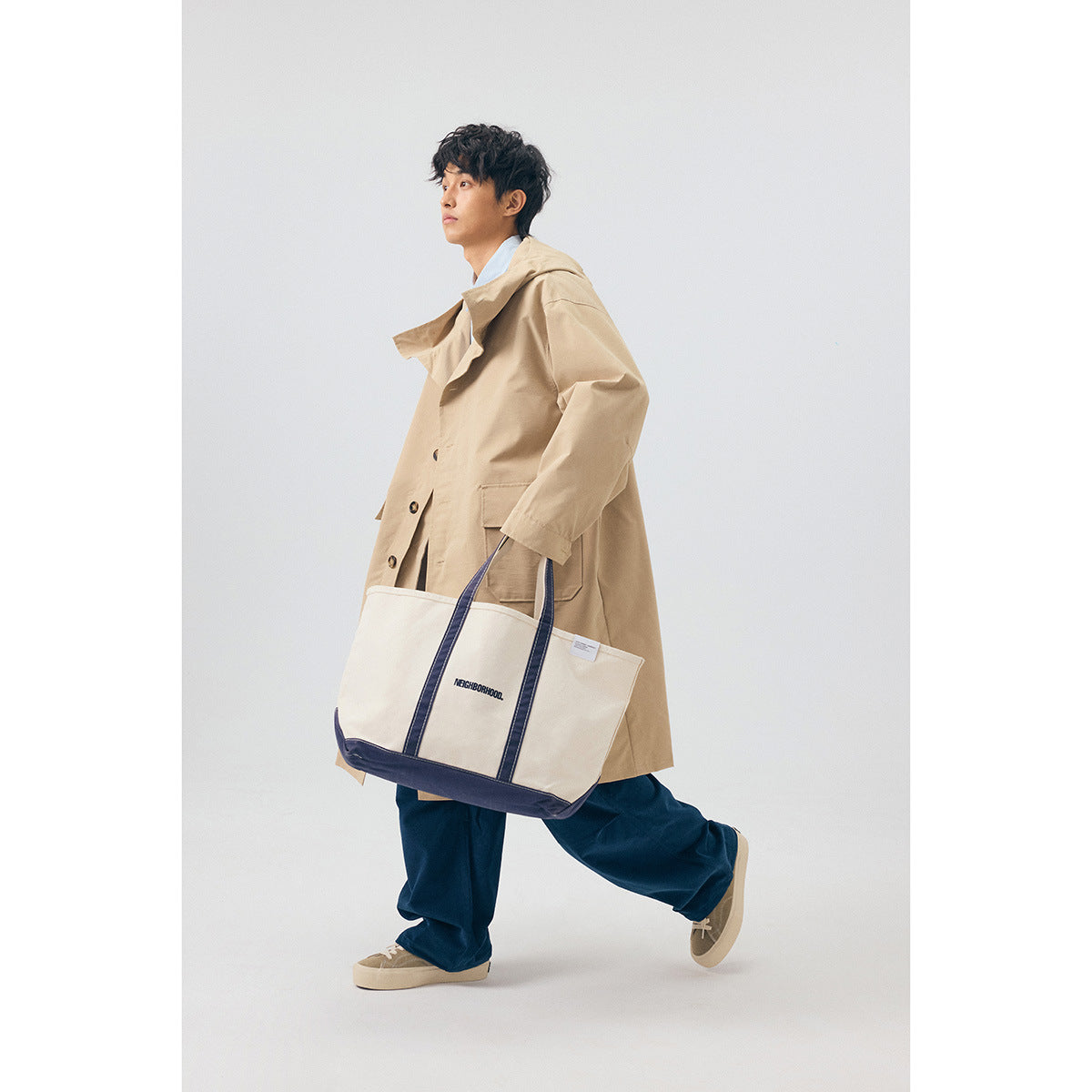 Model wearing a long beige hooded coat with navy trousers and carrying a cream tote with navy trim.