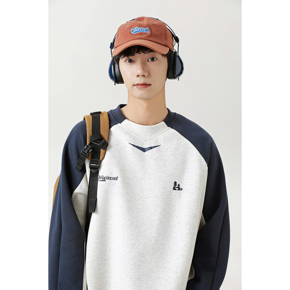 Man wearing a light gray sweatshirt with navy raglan sleeves, shown with a brown cap and backpack.