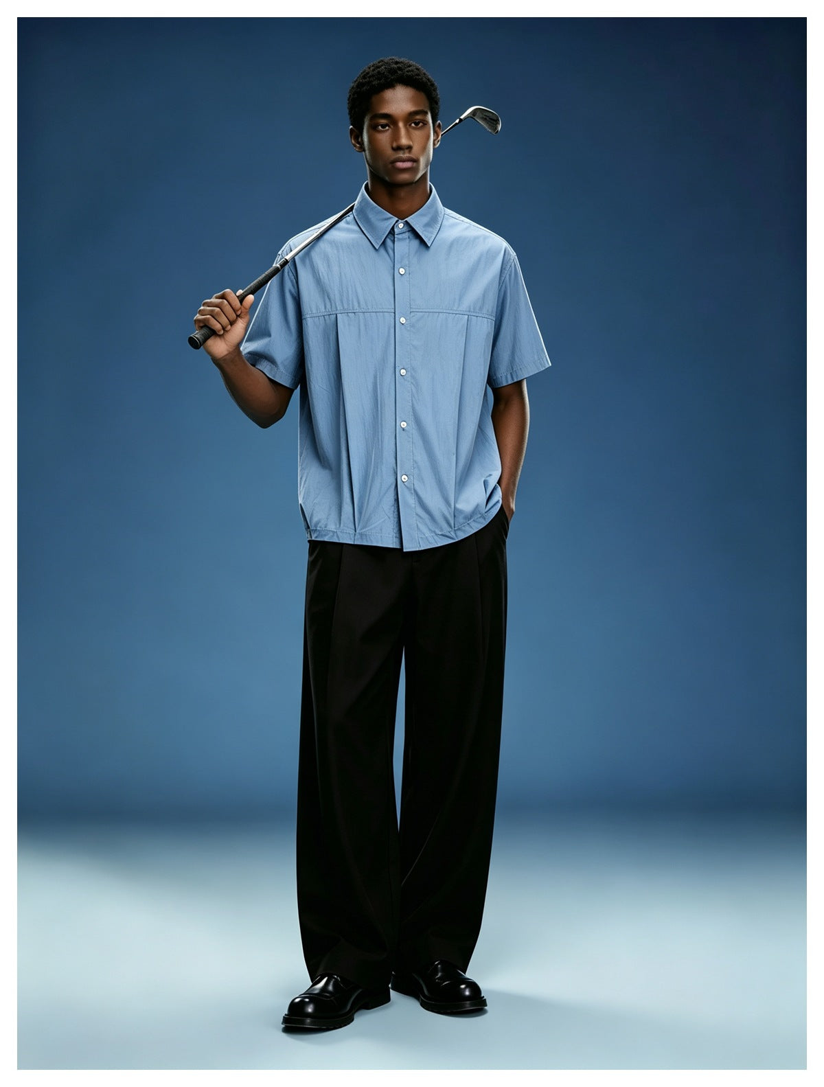 Model wearing a blue short-sleeve button-up shirt with black wide-leg trousers and black shoes against a blue backdrop