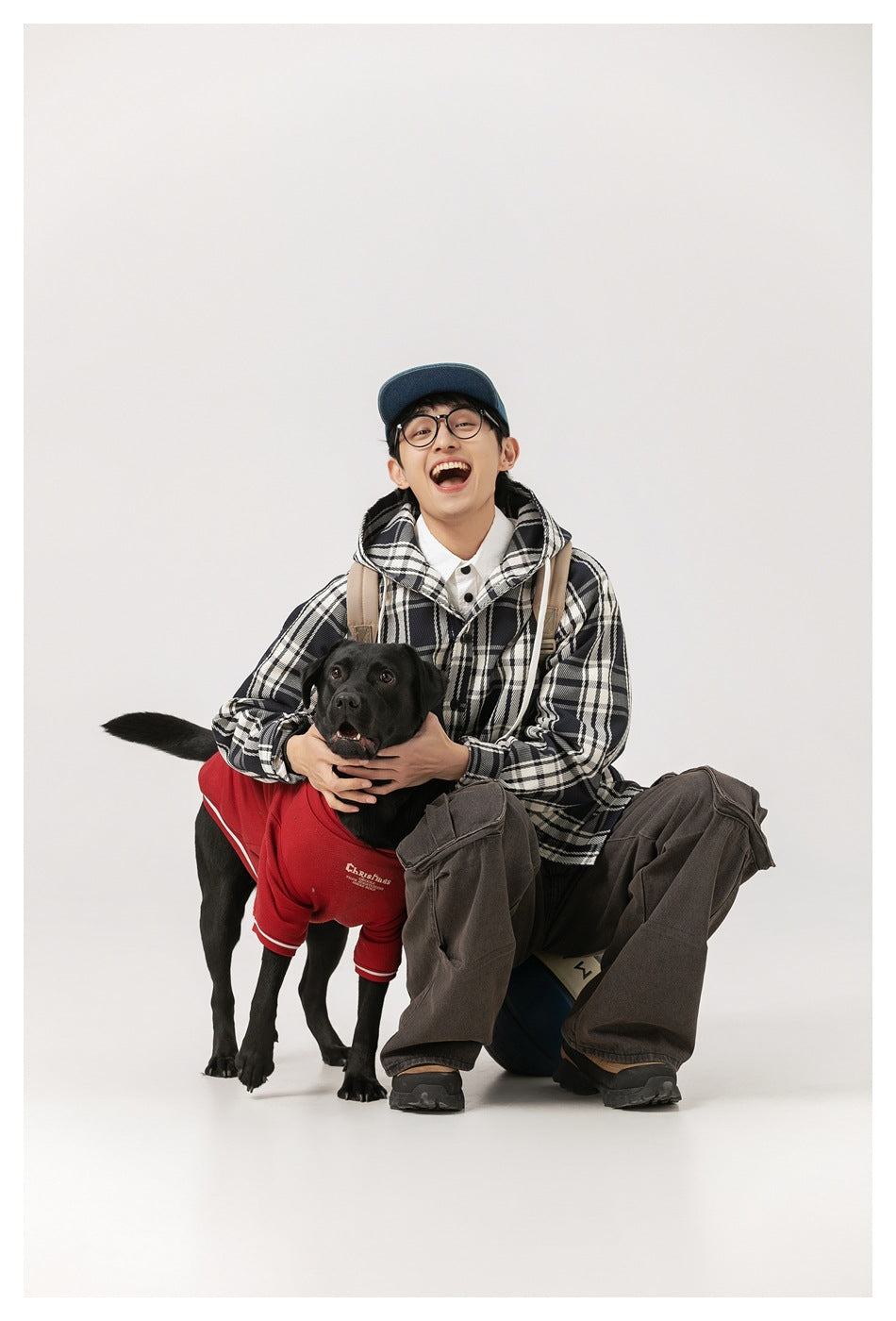 Model crouching in a black-and-white plaid hooded coat beside a black dog in a red sweater.