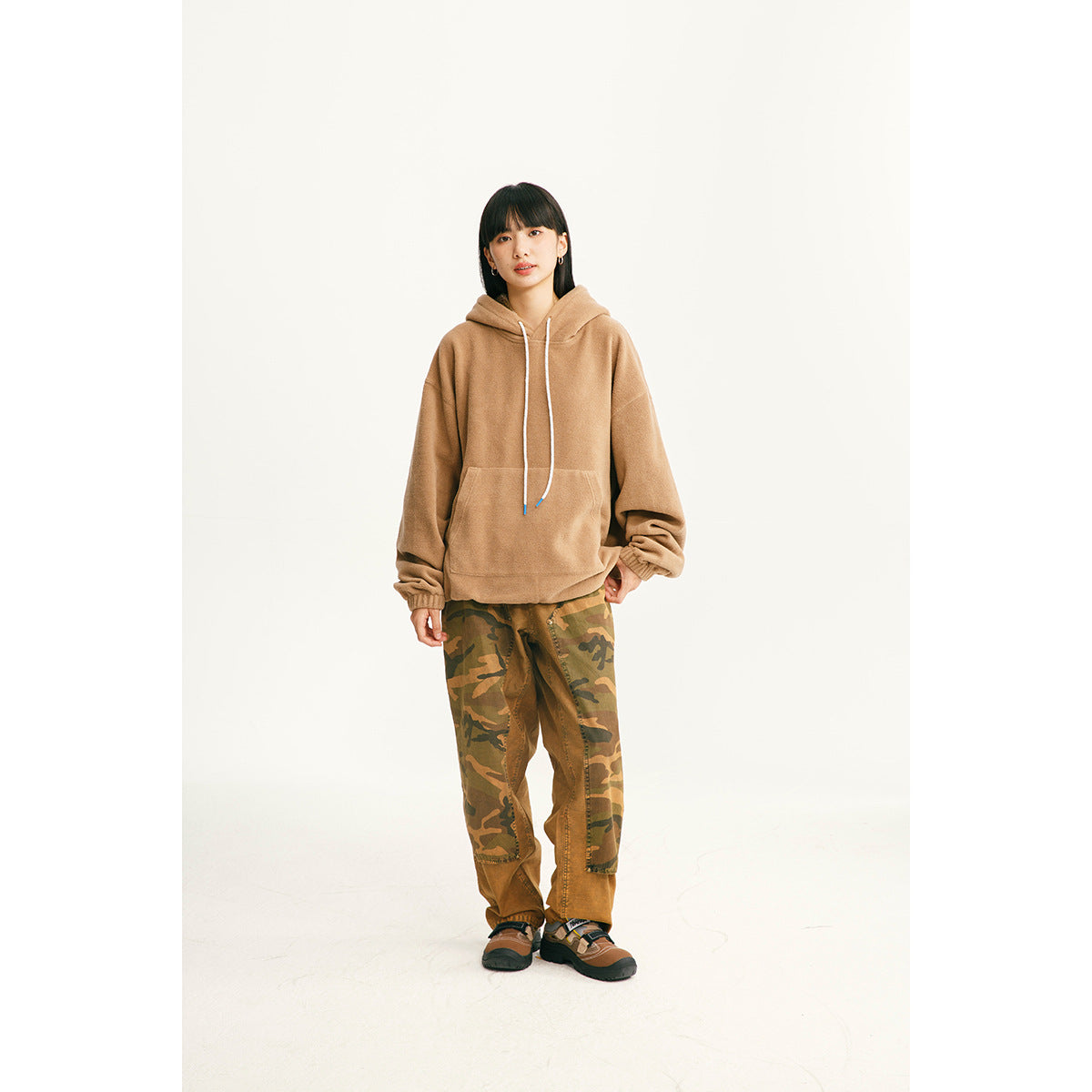 Model wearing a tan pullover hoodie with a kangaroo pocket and white drawstrings, paired with camouflage pants.