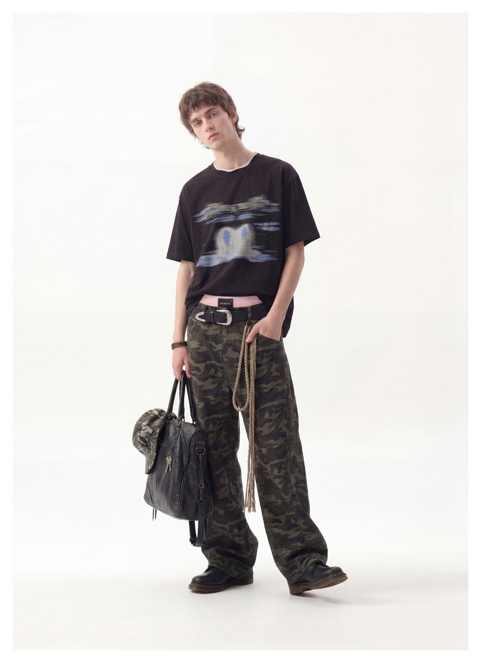 Man wearing a black graphic T-shirt with camouflage pants, holding a black bag and a camo hat, full-length view