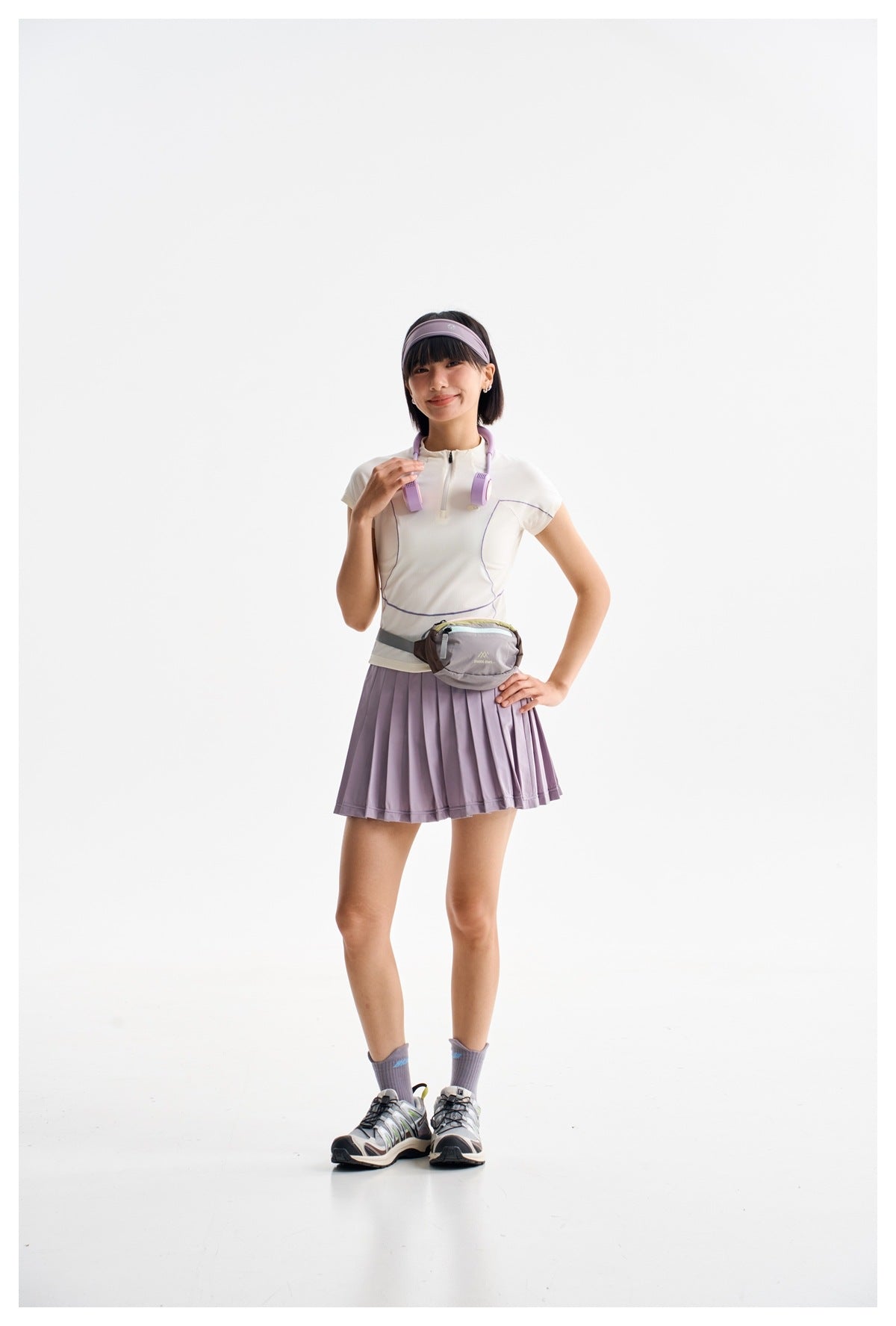 Model wearing an ivory quarter-zip top and lavender pleated skirt, with a lavender headband and gray waist bag.