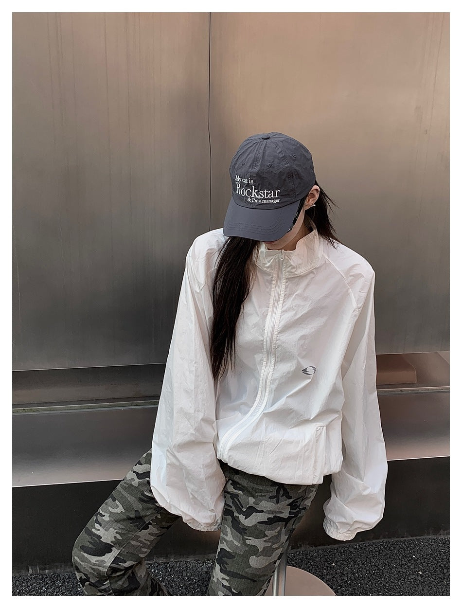 Model wearing a white zip-front jacket with a small chest emblem, paired with camouflage pants and a gray cap.