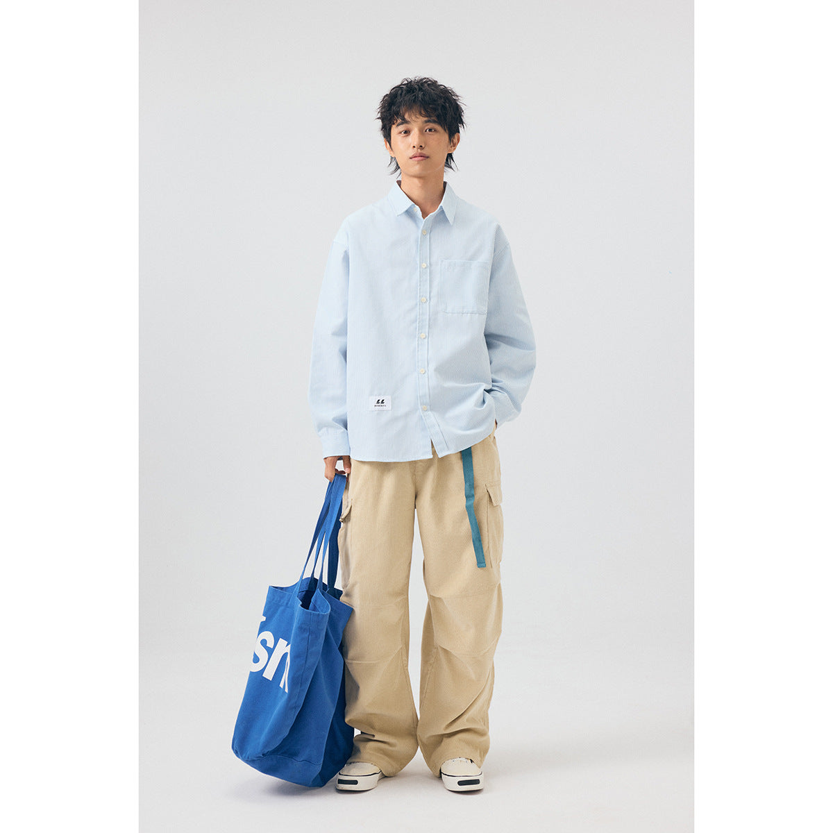 Model wearing a pale blue button-up shirt with a chest pocket and beige cargo pants, carrying a blue tote.