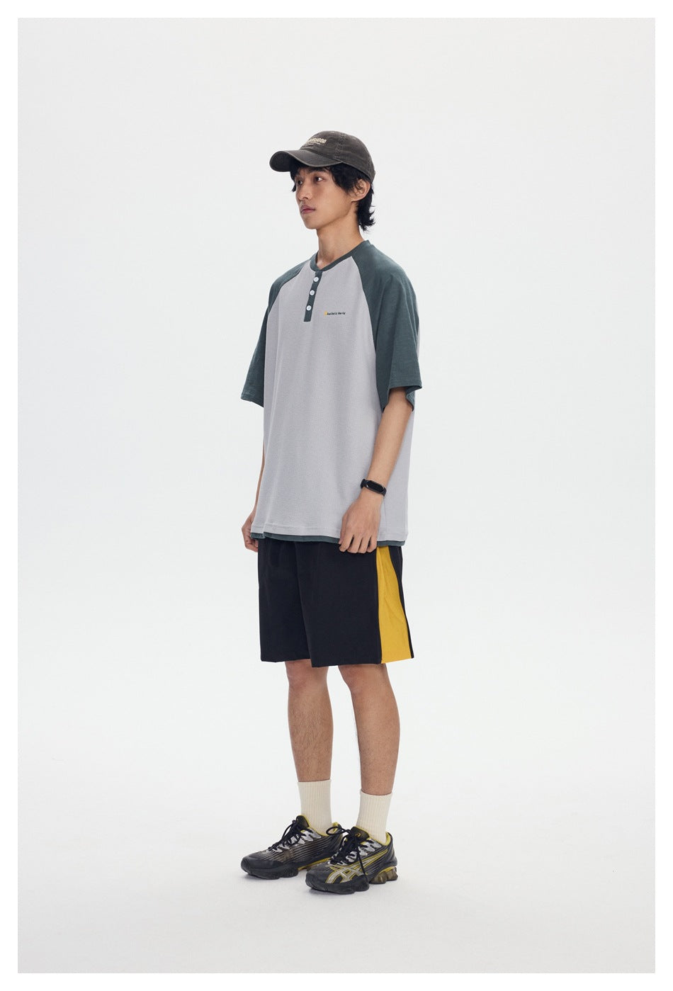 Model in a light gray Henley T-shirt with green raglan sleeves, black shorts with a yellow stripe, cap and sneakers, side