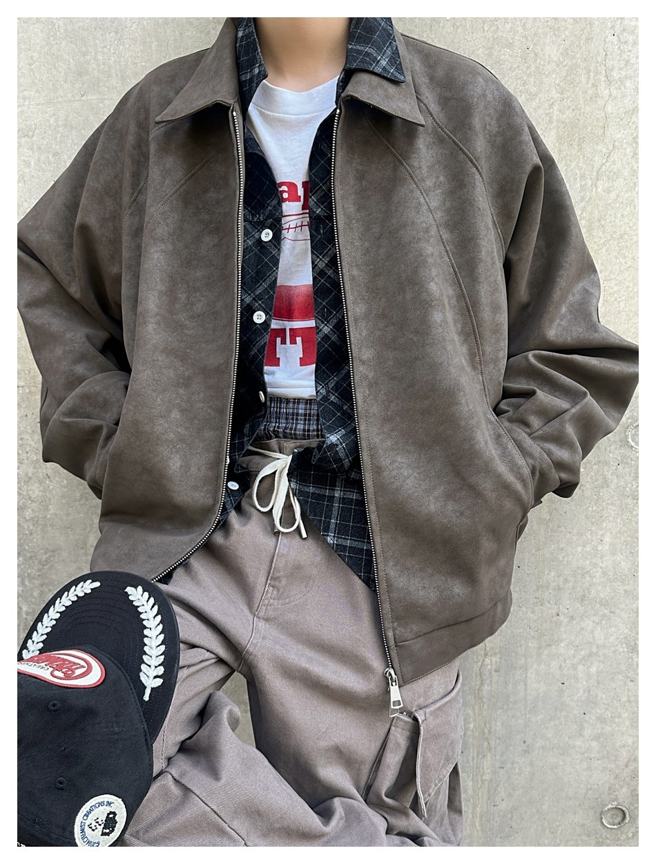 Close view of a person wearing an open brown faux suede jacket over a plaid shirt and graphic tee, holding a black cap