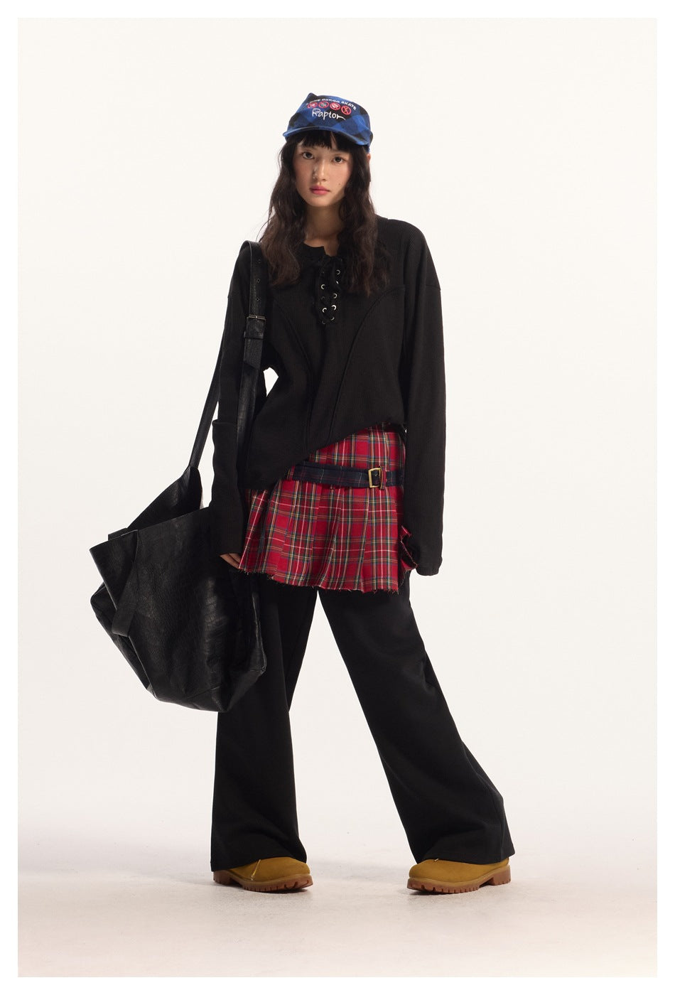 Model wearing black lace-up long-sleeve top with plaid pleated skirt, wide black pants, cap and large black bag