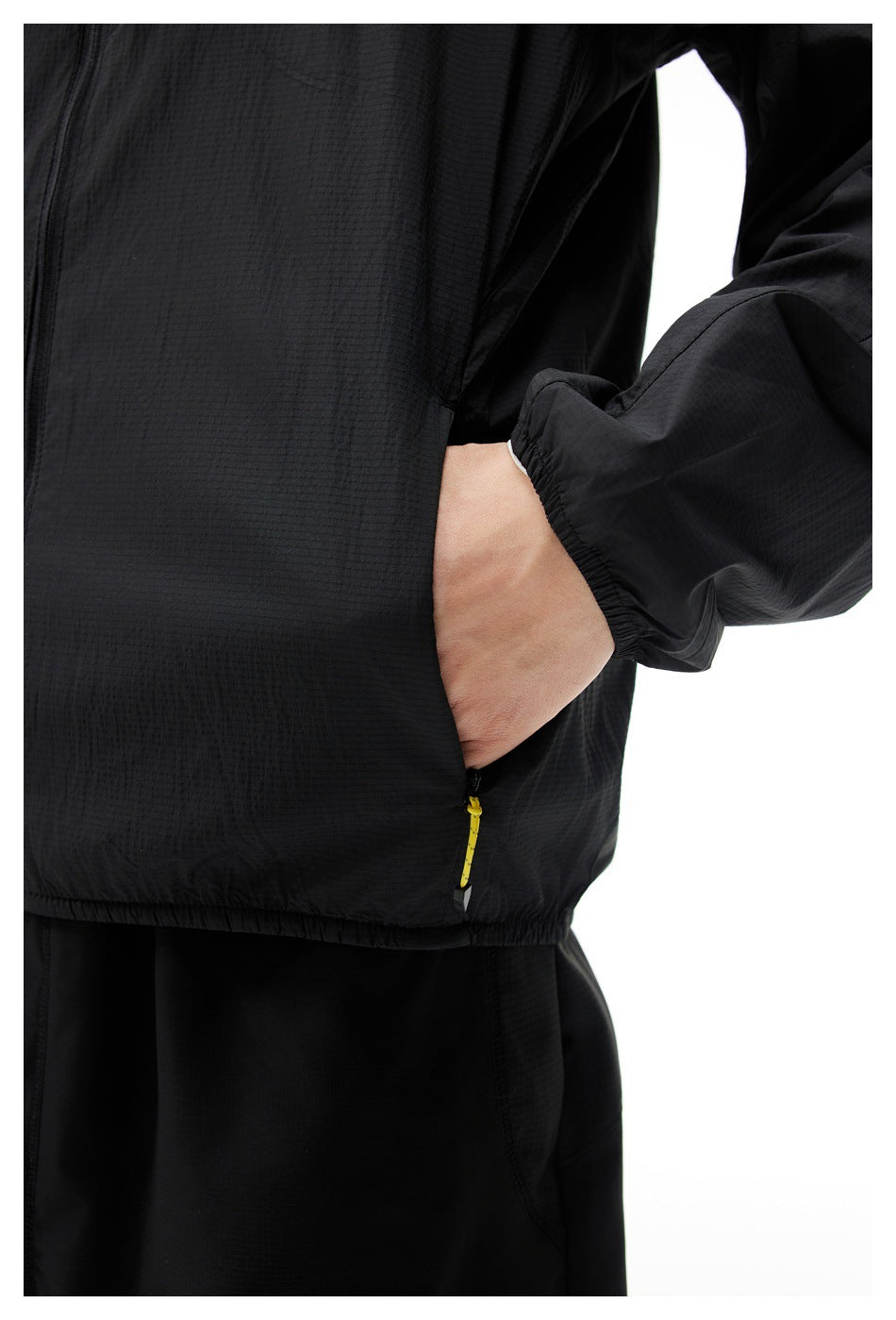 Close-up of a black jacket's textured front and elastic cuff, with a hand in the pocket and a yellow zipper pull.
