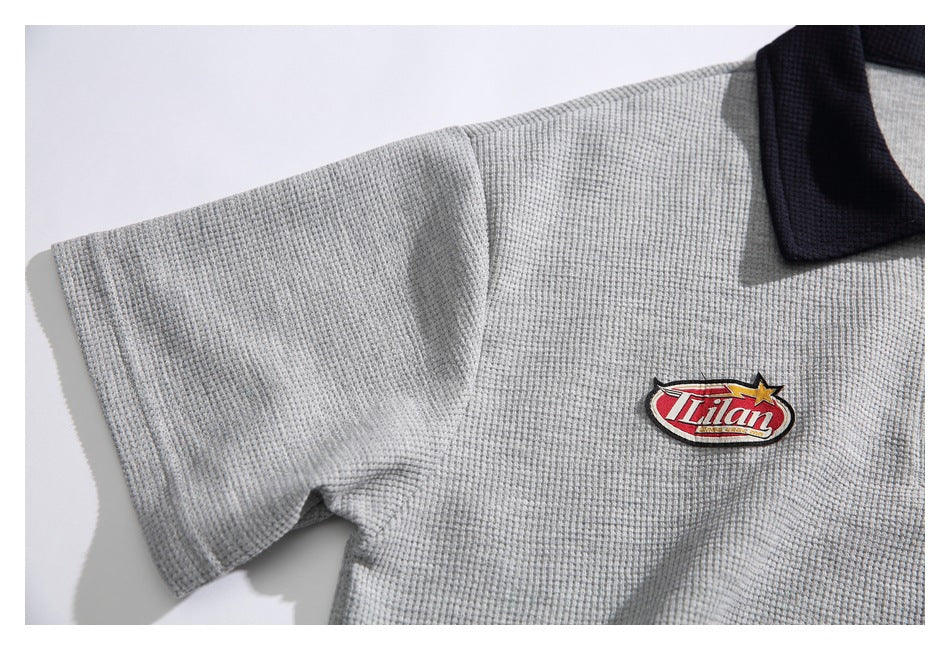 Close-up of a light gray textured polo shirt with a dark collar and an embroidered red-and-white chest emblem.