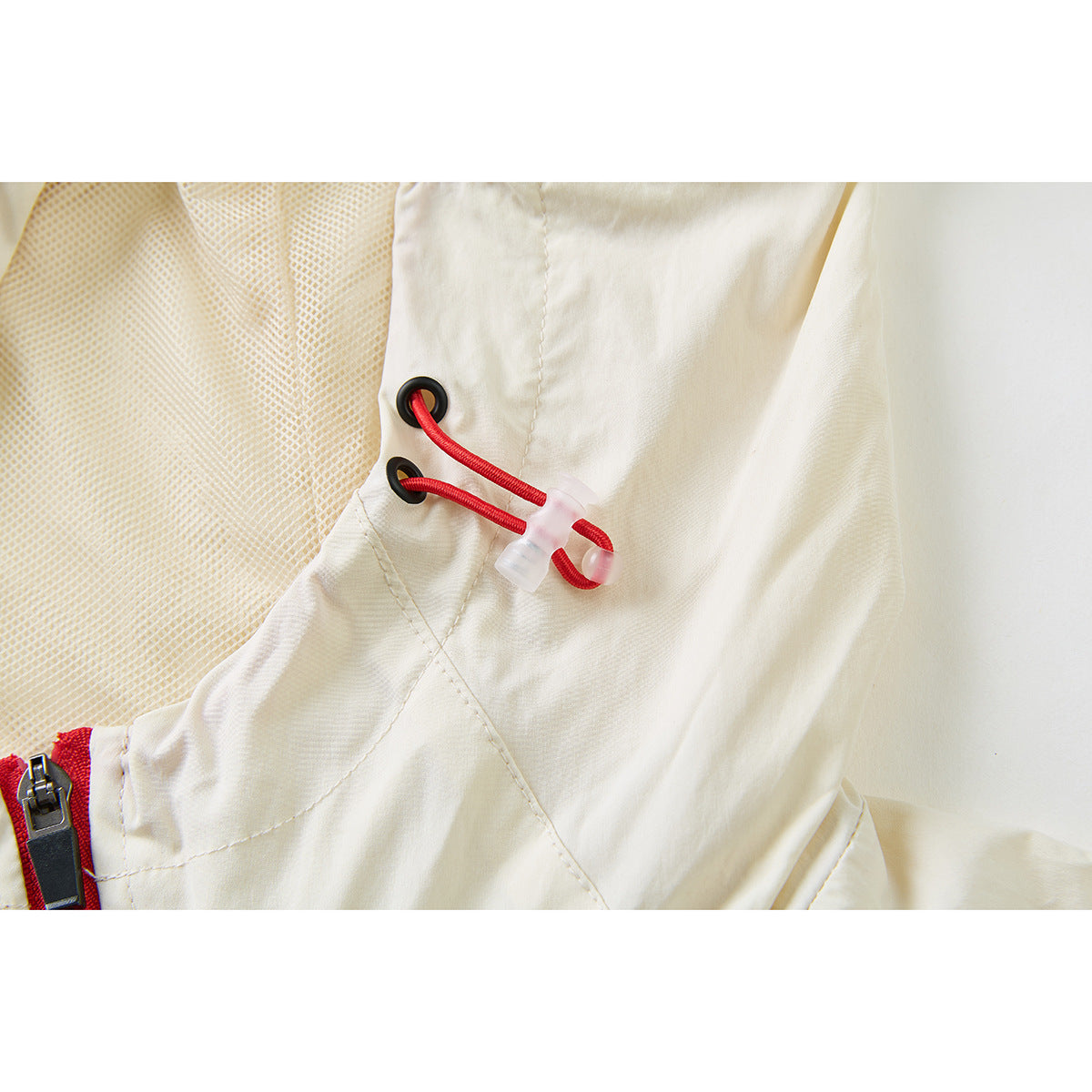 Close-up of a cream jacket hood with mesh lining, black eyelets, red drawcord and a red zipper edge.