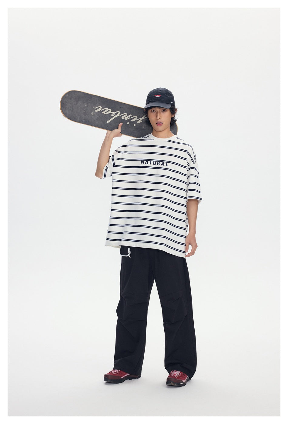 Model wearing an oversized white T-shirt with thin navy stripes and NATURAL text, black pants, and a cap, holding a