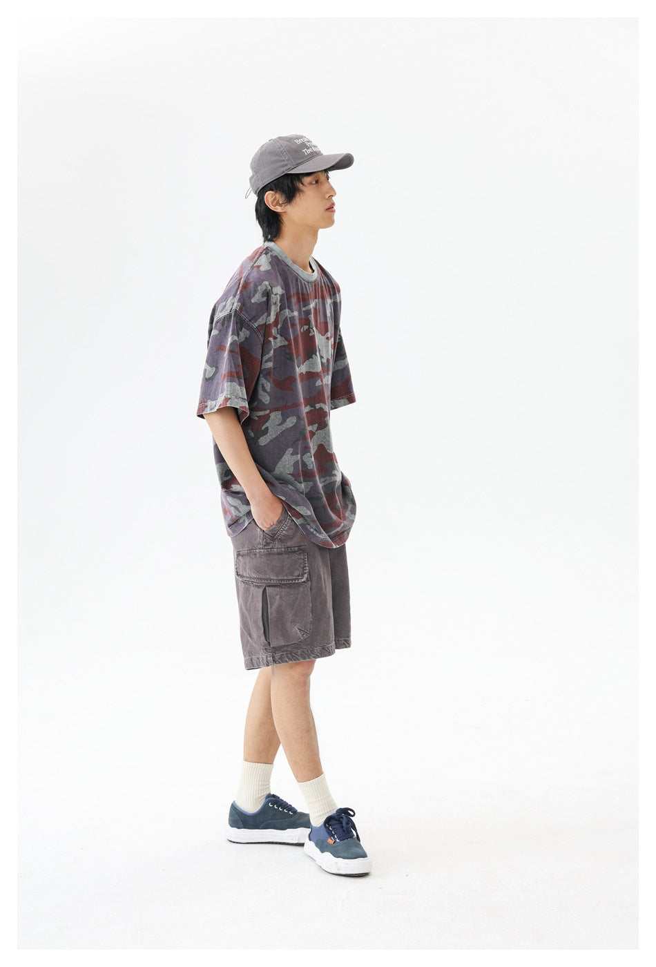 Side view of a man in an oversized camouflage T-shirt, brown cargo shorts, gray cap and navy sneakers