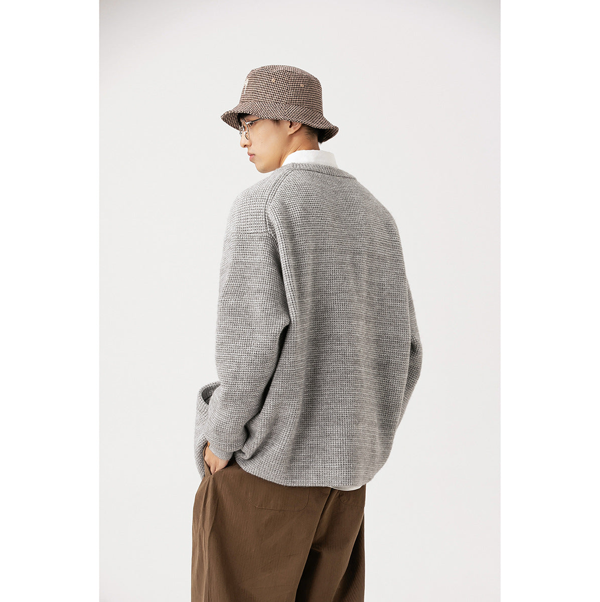 Rear view of a model wearing a light gray textured cardigan with brown trousers and a brown checked bucket hat.