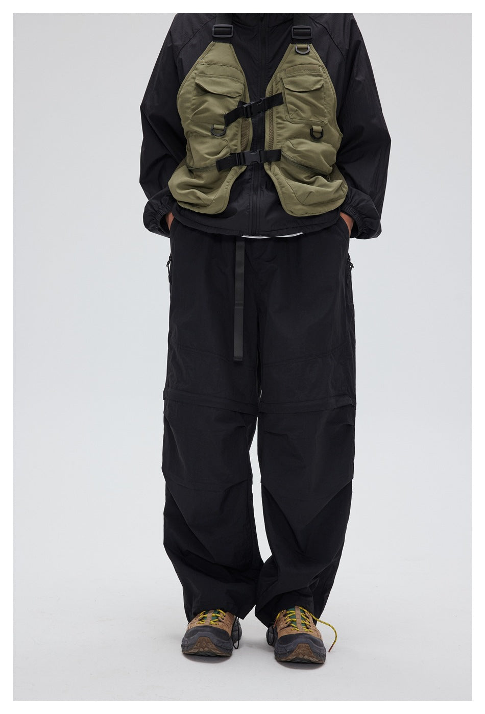 Front view of a model wearing loose black pants with zip pockets, layered panels, and an olive utility vest.