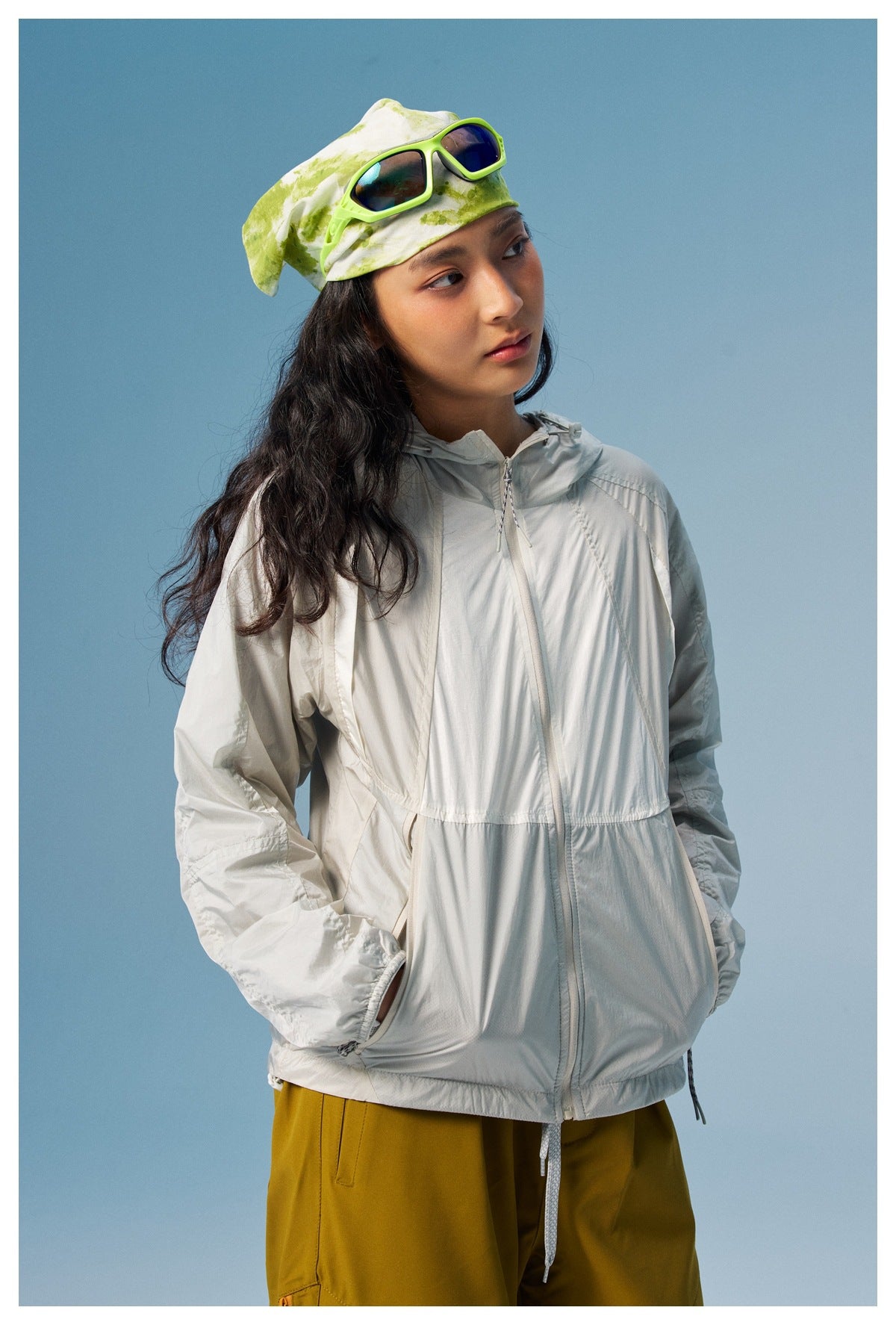 Woman wearing a light gray hooded zip jacket and mustard pants, with a green-and-white patterned headscarf.