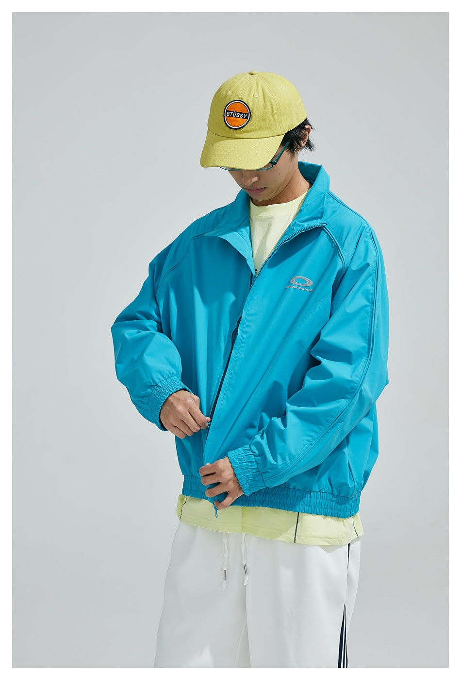 Person zipping a sky blue jacket with raglan sleeves and elastic hem, wearing a yellow cap and white pants