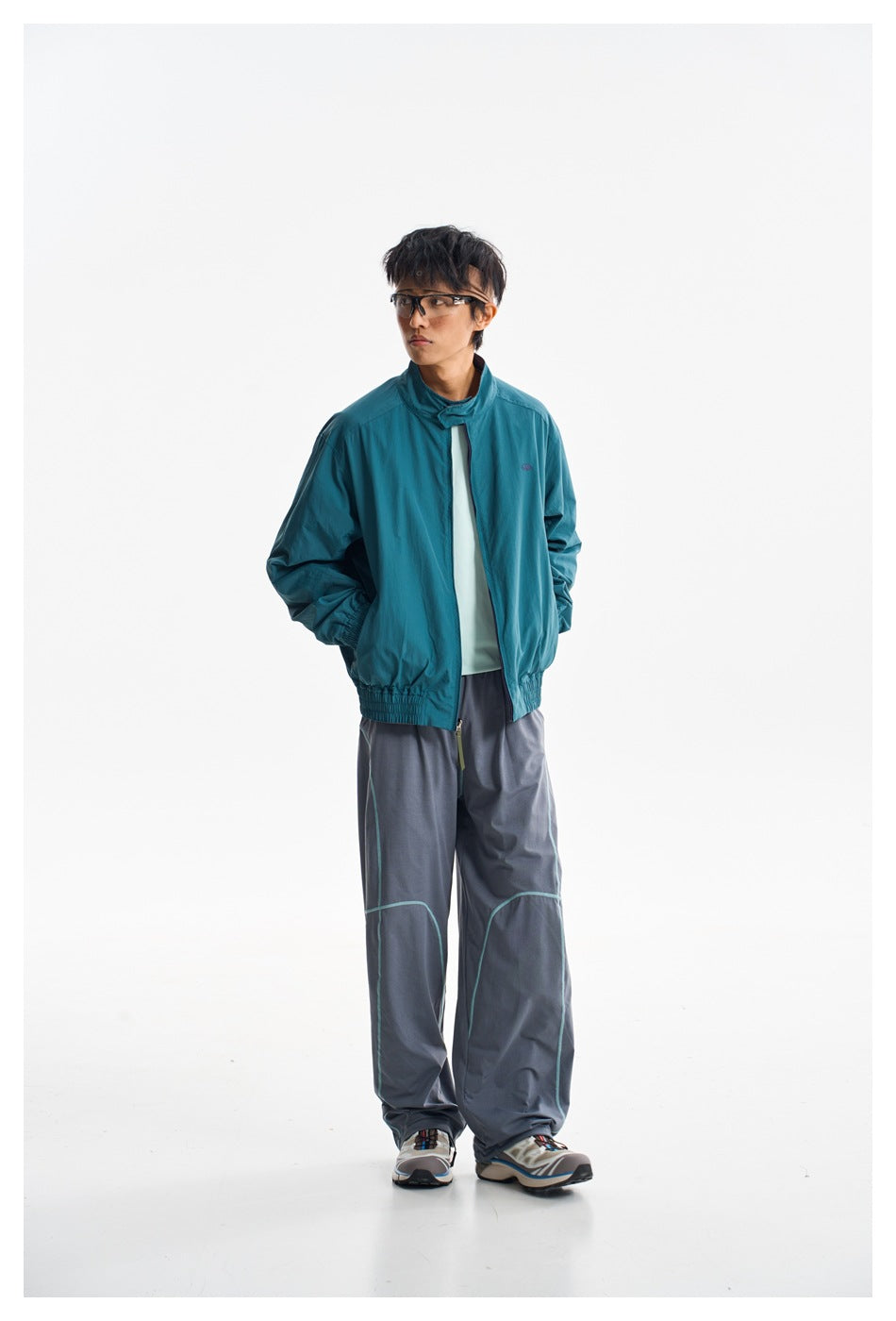 Full-length model wearing a teal zip jacket and loose gray trousers with curved turquoise piping.