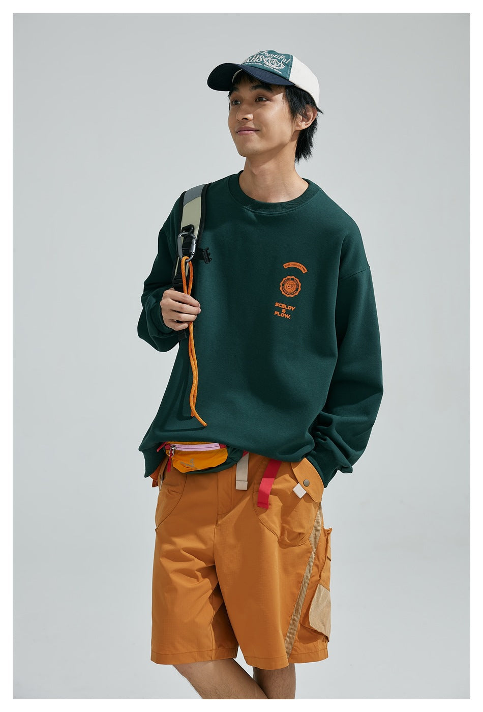 Model wearing a dark green crewneck sweatshirt with an orange chest graphic and mustard cargo shorts, shown at an angle.