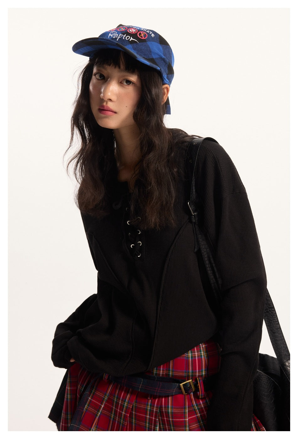 Closer view of a model in an oversized black ribbed lace-up top and red plaid skirt with a blue cap.