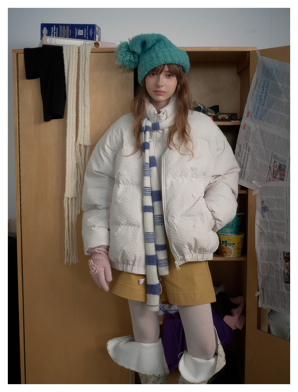 Model faces forward in a white quilted jacket, blue-and-white scarf, teal hat and mustard shorts.