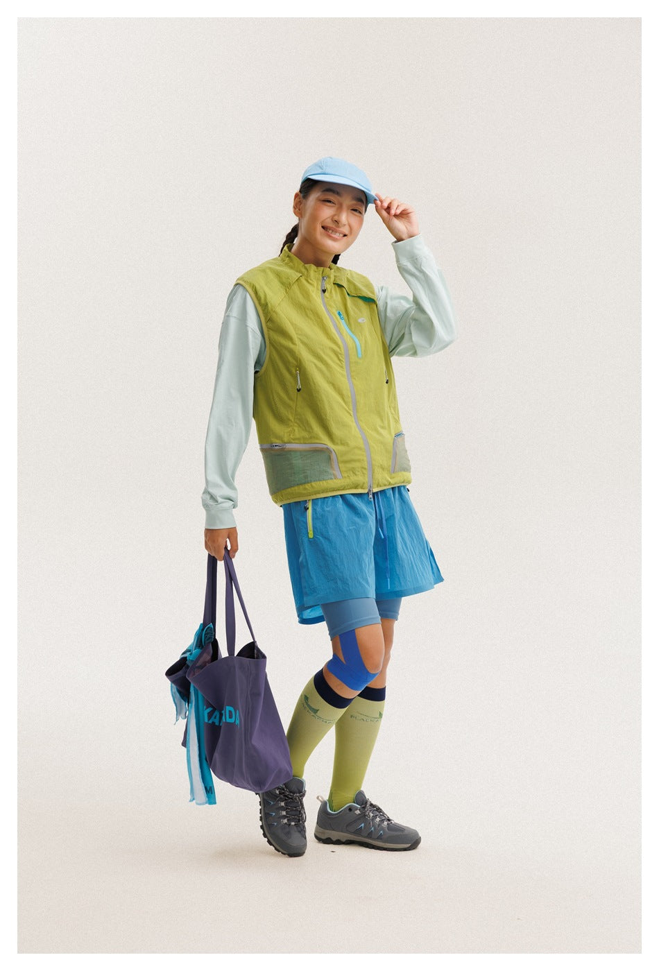 A model shows the front of a lime-green zip vest with a pale mint top and blue shorts, carrying a purple tote.