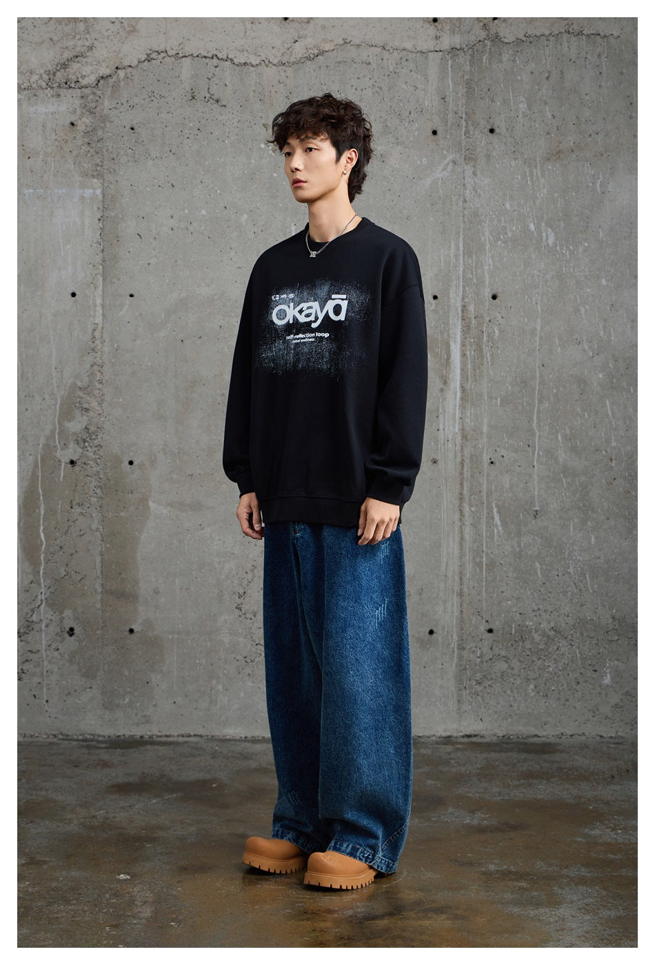 Full-length model wearing a black graphic crewneck sweatshirt, wide blue jeans, and tan shoes.