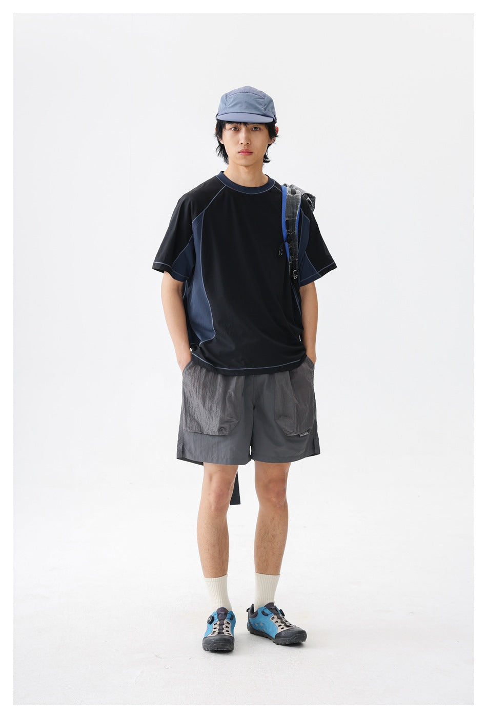 Front view of a man wearing a black and navy color-block T-shirt, gray utility shorts and a blue-gray cap, with a backpack.