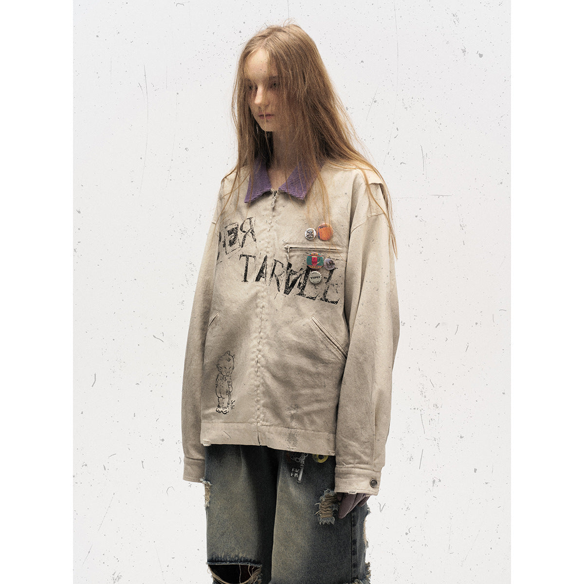 Model wearing a beige graphic jacket with a purple collar, chest pins and oversized dark distressed denim shorts.