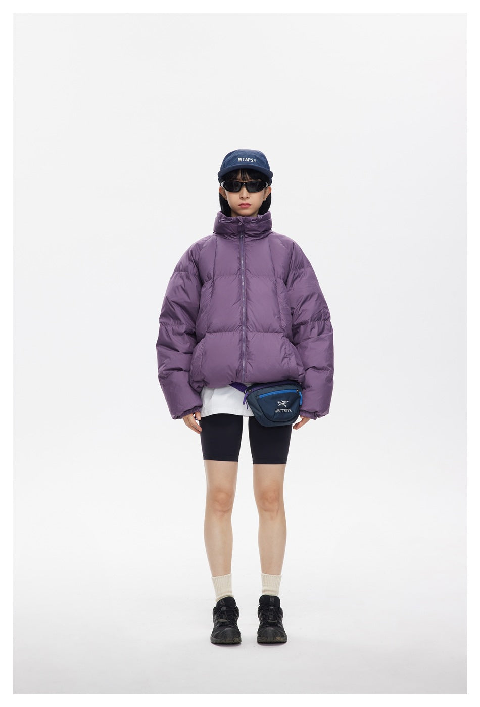 Model wearing a purple quilted puffer jacket with a high collar, black shorts, cap, and crossbody bag.