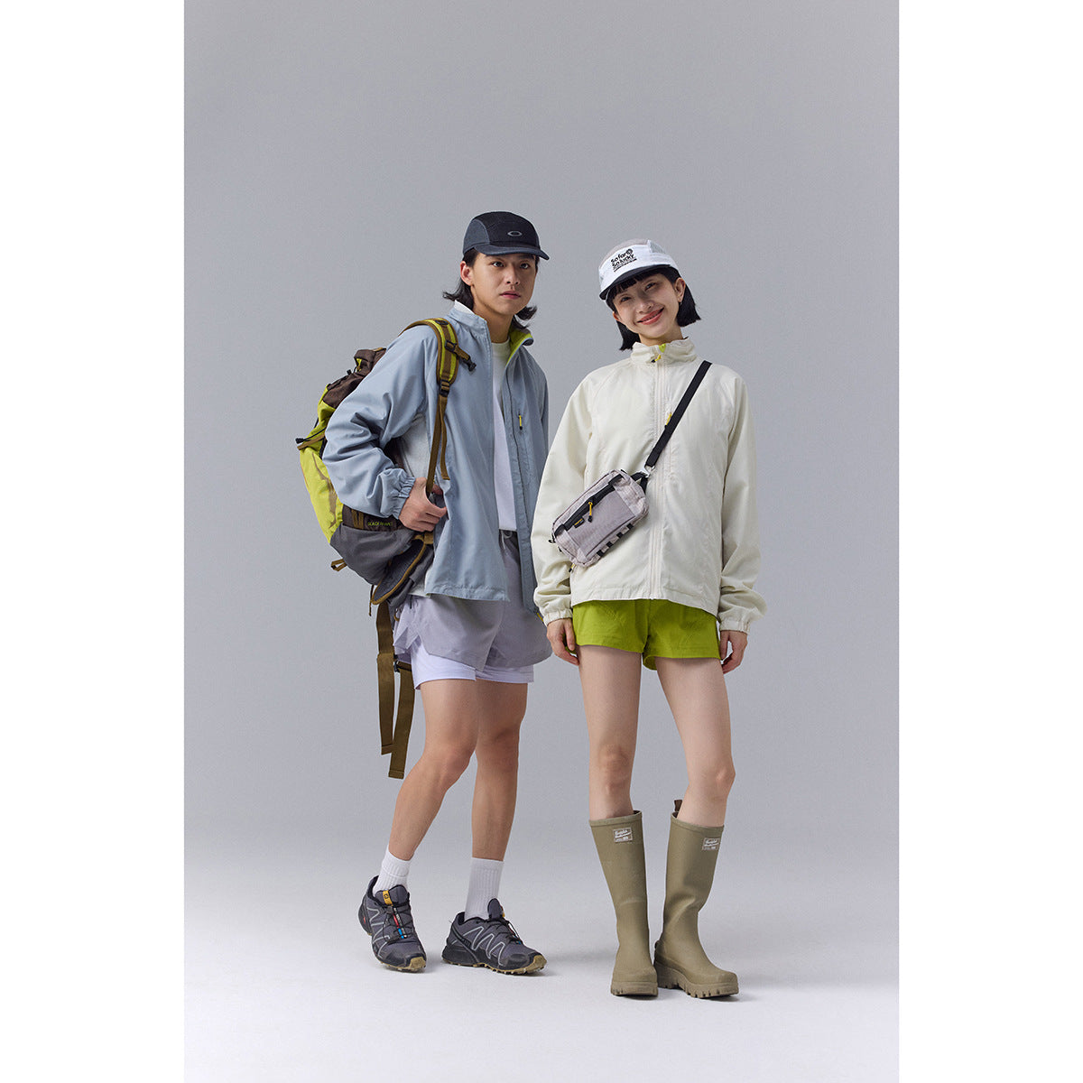 Two models wear outdoor jackets, bright shorts, hiking footwear and accessories; one jacket is pale blue and the other cream.
