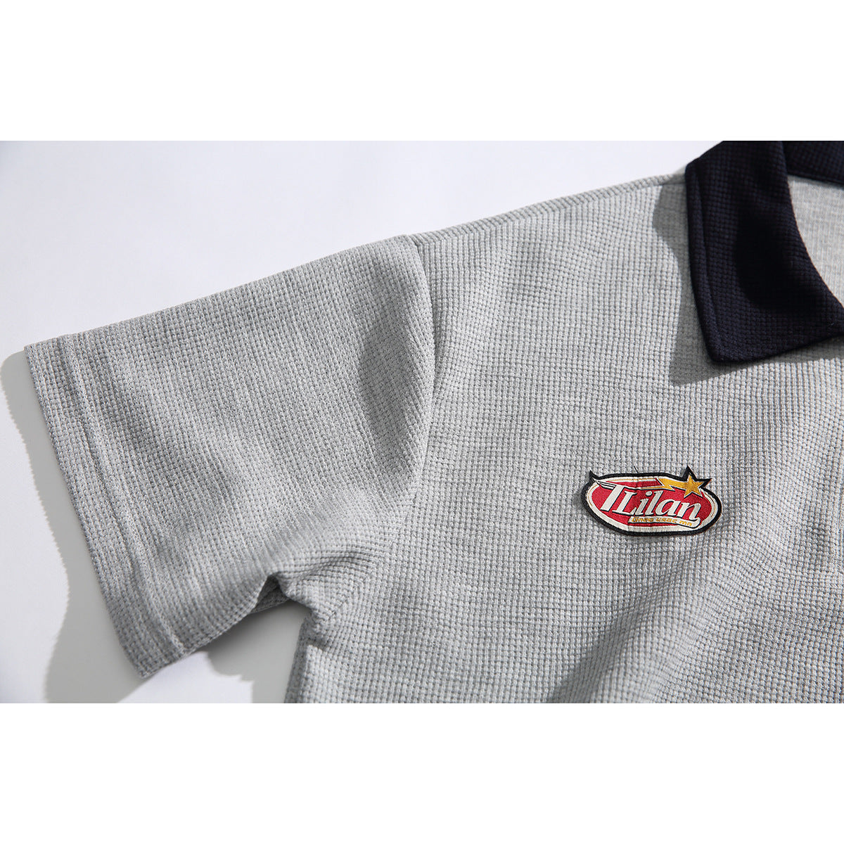 Close-up of the gray polo’s textured sleeve and chest, showing a red embroidered patch and dark collar.