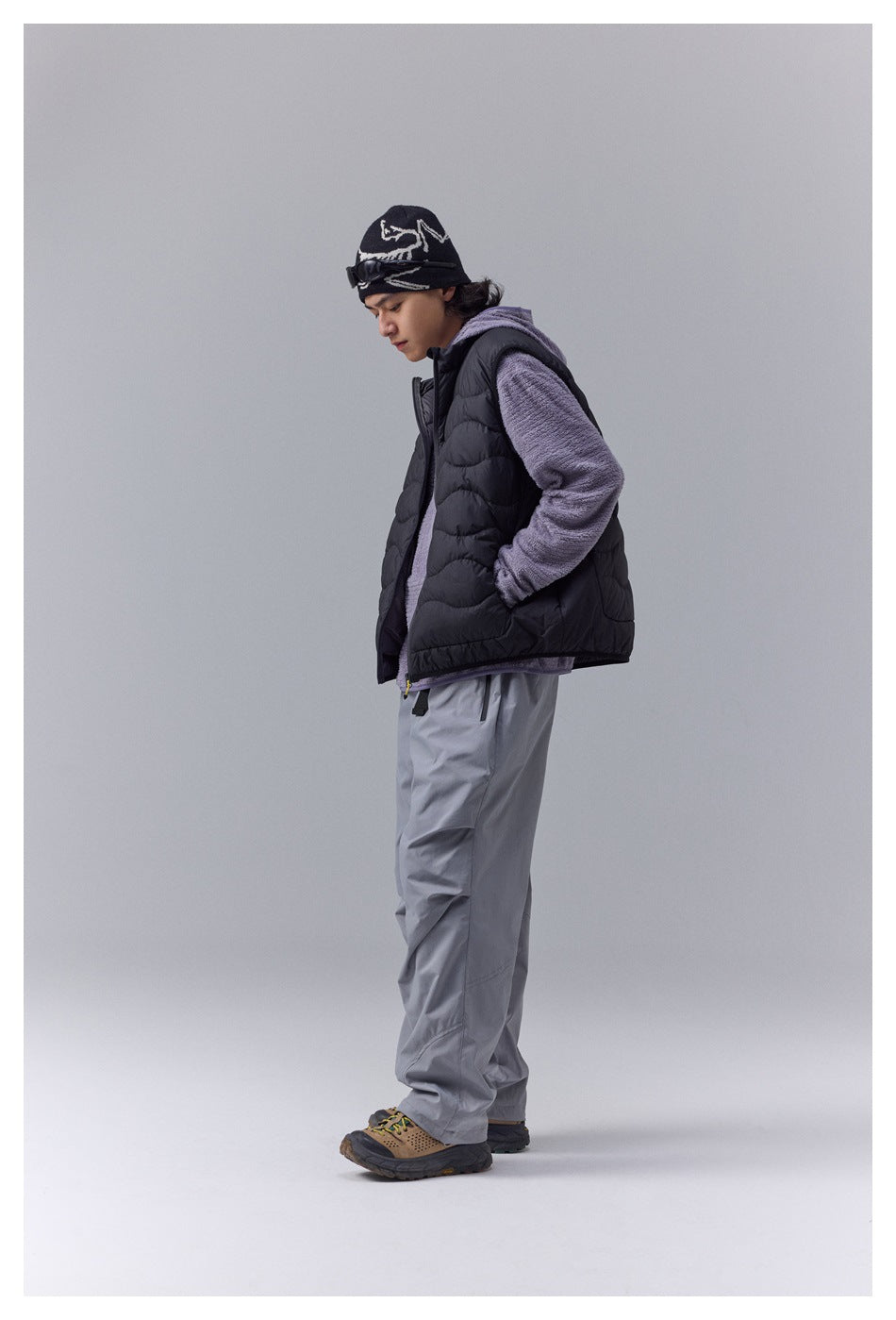 Side view of a model in a black quilted vest over a purple textured top, hand in pocket, with gray pants