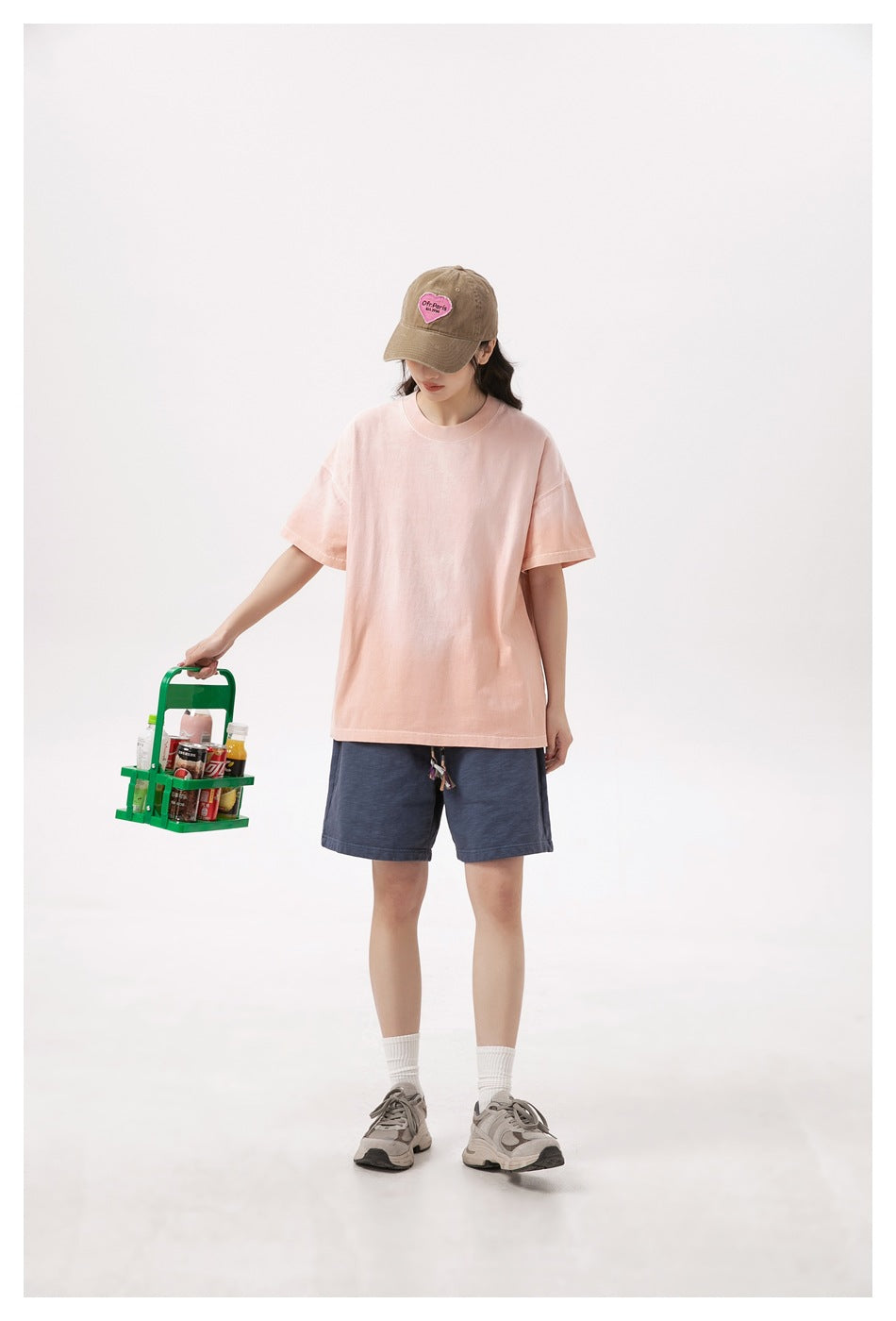 Model wearing a pink oversized T-shirt with navy shorts, a brown cap and sneakers, holding a green drink carrier