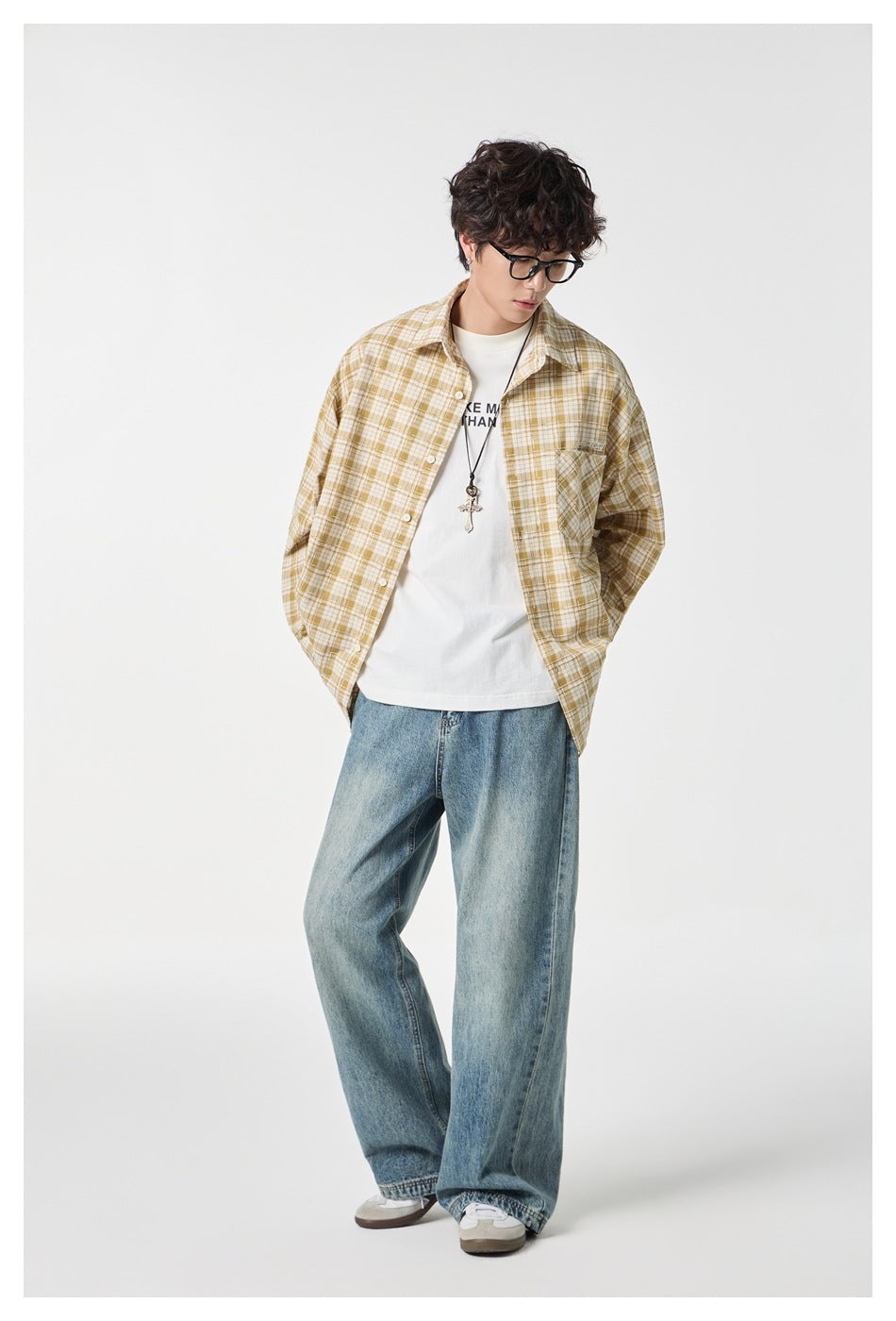 Model looking down in an open mustard and cream plaid button-up shirt, white tee, and loose blue jeans.