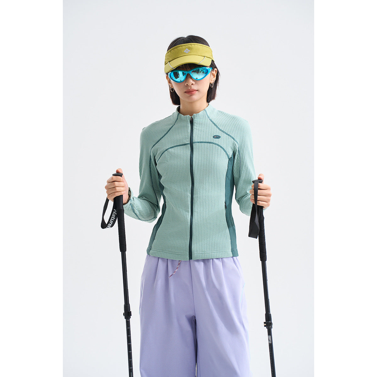 Model wearing a textured mint-green zip jacket with dark teal panels and lavender trousers, holding trekking poles.