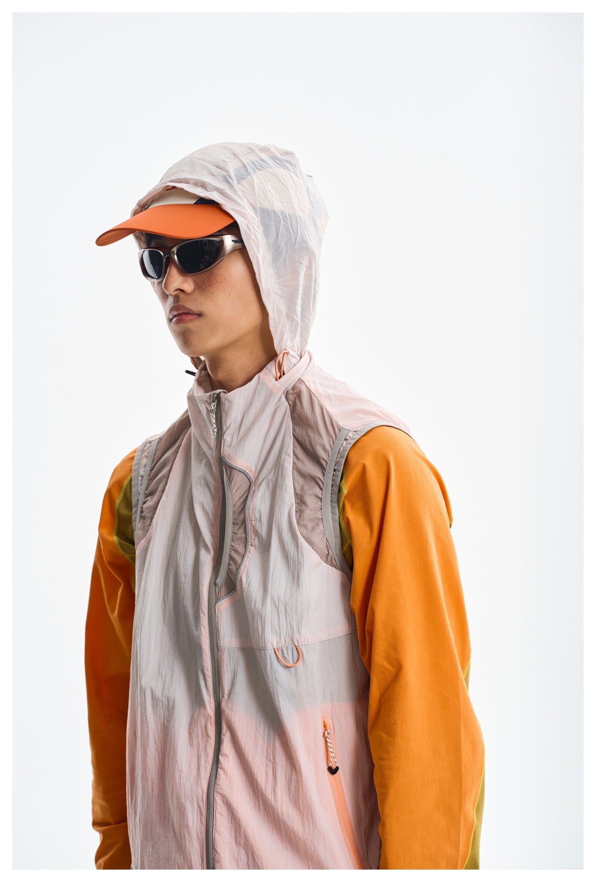 Model wearing a pale blush hooded vest over an orange long-sleeve top, with an orange cap and dark sunglasses.
